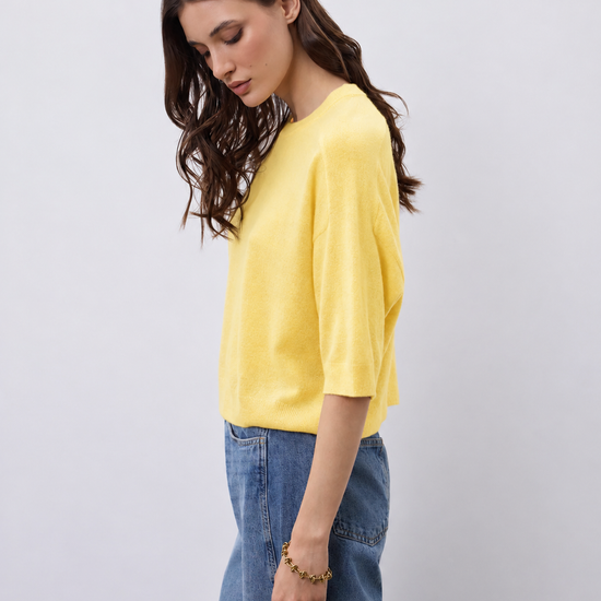 A woman wearing a bright yellow short-sleeve knit top and blue jeans, standing against a light gray studio background.