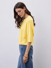 A woman wearing a bright yellow short-sleeve knit top and blue jeans, standing against a light gray studio background.