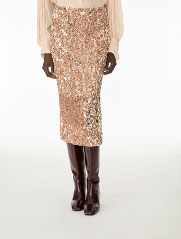 A model is shown from the waist down wearing a rose gold sequined midi skirt, a cream blouse, and dark brown knee-high boots against a white studio background.