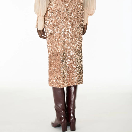 A model shown from the back wearing a high-waisted, knee-length sequined midi skirt in a champagne rose gold tone, paired with a light beige blouse and dark brown knee-high boots, against a plain white studio background.