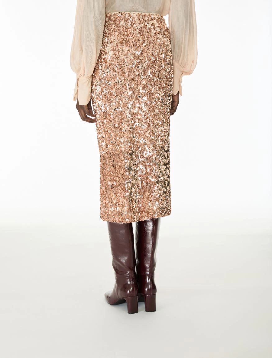 A model shown from the back wearing a high-waisted, knee-length sequined midi skirt in a champagne rose gold tone, paired with a light beige blouse and dark brown knee-high boots, against a plain white studio background.