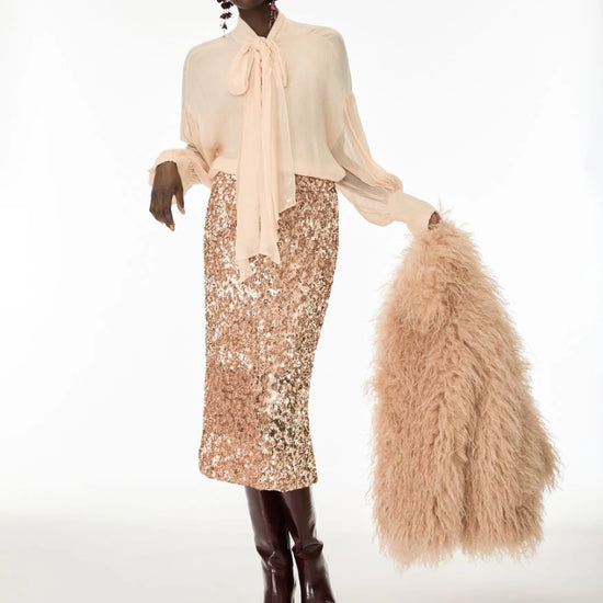 A model stands against a white studio background wearing a pale blush long-sleeve blouse with a large bow tie-neck, a champagne sequined midi skirt, dark brown knee-high boots, and holding a fluffy blush faux-fur accessory.