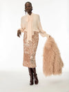 A model stands against a white studio background wearing a pale blush long-sleeve blouse with a large bow tie-neck, a champagne sequined midi skirt, dark brown knee-high boots, and holding a fluffy blush faux-fur accessory.