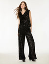 A woman modeling a black sequined sleeveless jumpsuit with a draped neckline and wide-leg pants, photographed against a plain white studio background.