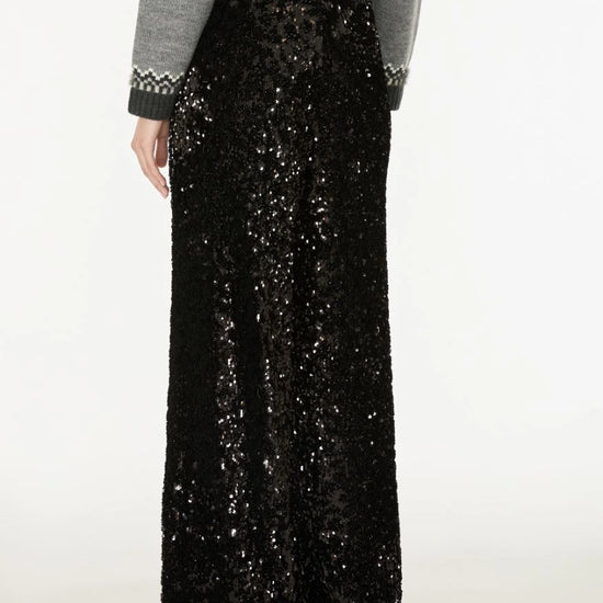 A person wearing a long black sequined maxi skirt with a gray long-sleeve top featuring a black and white patterned knit cuff, shown from the back against a plain white studio background.