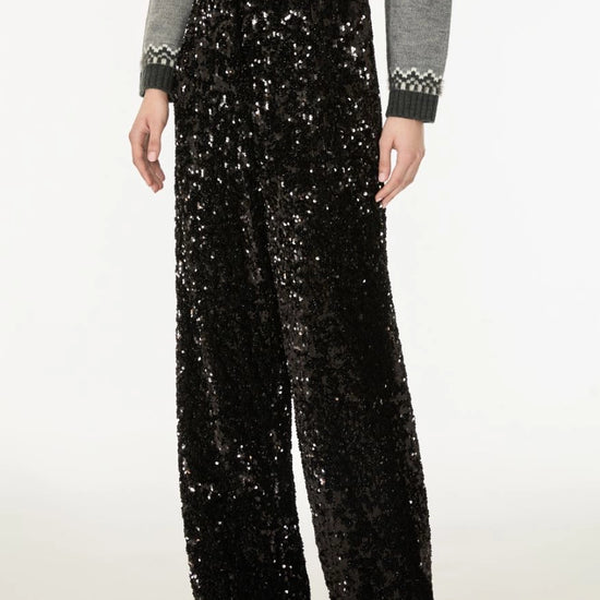 A model is shown from the waist down wearing high-waisted, wide-leg black sequined trousers paired with a gray cropped sweater and black shoes, set against a plain white studio background.