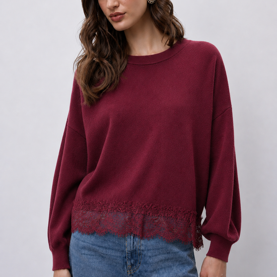 A woman wearing a long-sleeve crewneck sweater in deep burgundy with a lace hem detail, paired with blue jeans, against a light gray studio background.