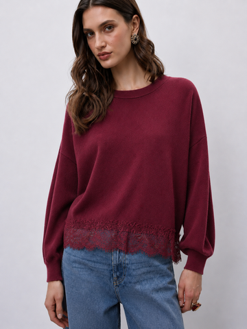 A woman wearing a long-sleeve crewneck sweater in deep burgundy with a lace hem detail, paired with blue jeans, against a light gray studio background.
