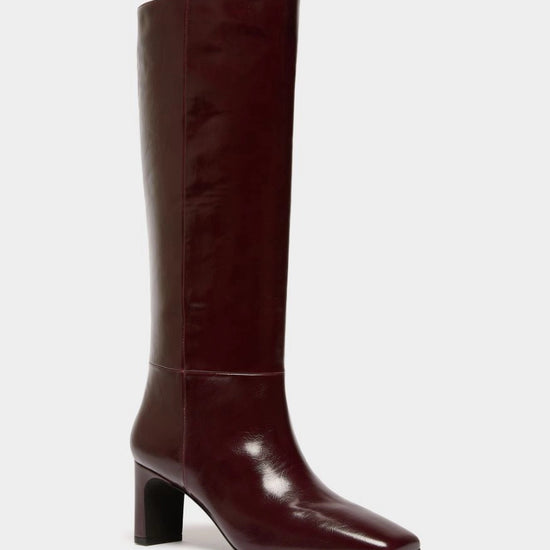 A single knee-high boot in a glossy deep burgundy leather, featuring a pointed toe and a chunky block heel, shown against a plain light gray background.