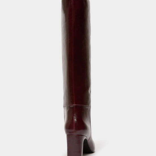 A single knee-high boot in a glossy deep burgundy leather, shown from a rear three-quarter view against a solid off-white background.