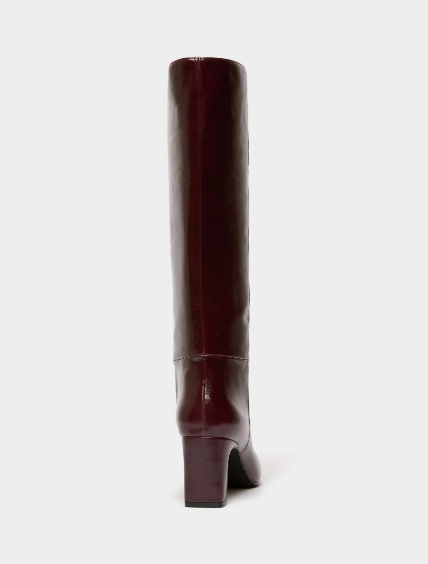 A single knee-high boot in a glossy deep burgundy leather, shown from a rear three-quarter view against a solid off-white background.