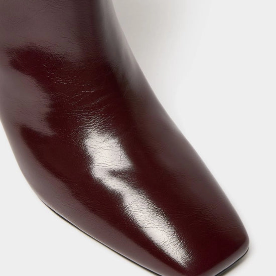 Close-up of a glossy dark burgundy leather boot showing the pointed toe and smooth texture against a light gray background.