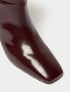 Close-up of a glossy dark burgundy leather boot showing the pointed toe and smooth texture against a light gray background.