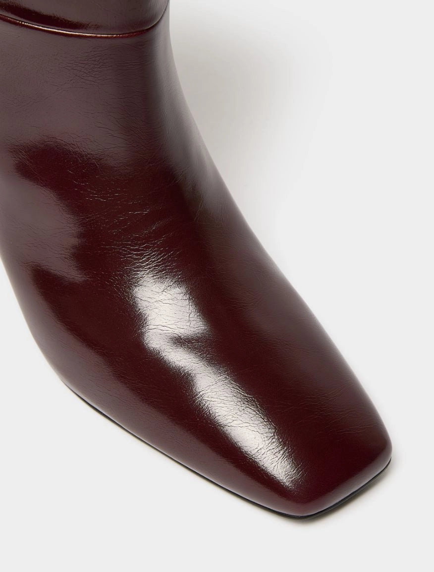 Close-up of a glossy dark burgundy leather boot showing the pointed toe and smooth texture against a light gray background.
