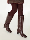 A pair of knee-high glossy dark brown boots worn with matching dark brown tights, shown against a plain light gray studio background.