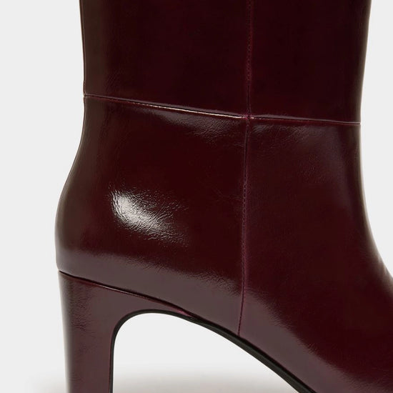 Close-up of a dark burgundy leather boot with a block heel, showing the side profile and glossy finish against a light gray background.