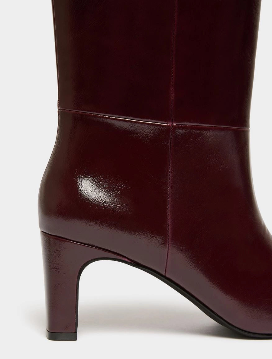 Close-up of a dark burgundy leather boot with a block heel, showing the side profile and glossy finish against a light gray background.