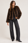 A woman modeling a dark brown fur jacket, styled with black wide-leg jeans and black pointed-toe heels, against a plain light gray studio background.