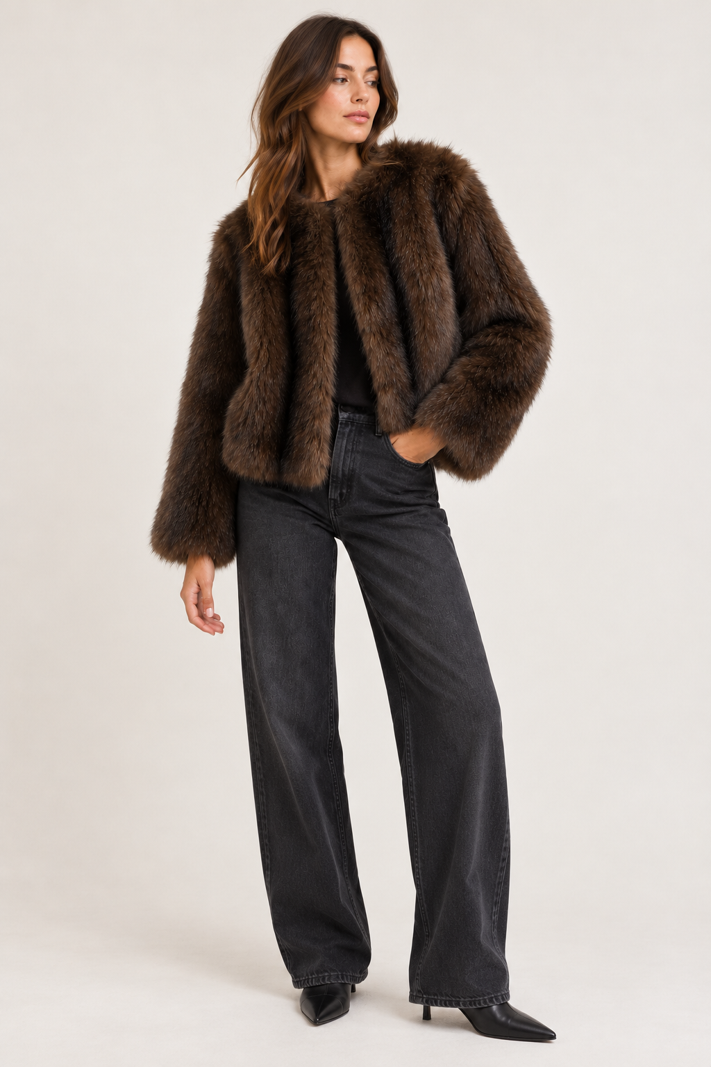 A woman modeling a dark brown fur jacket, styled with black wide-leg jeans and black pointed-toe heels, against a plain light gray studio background.