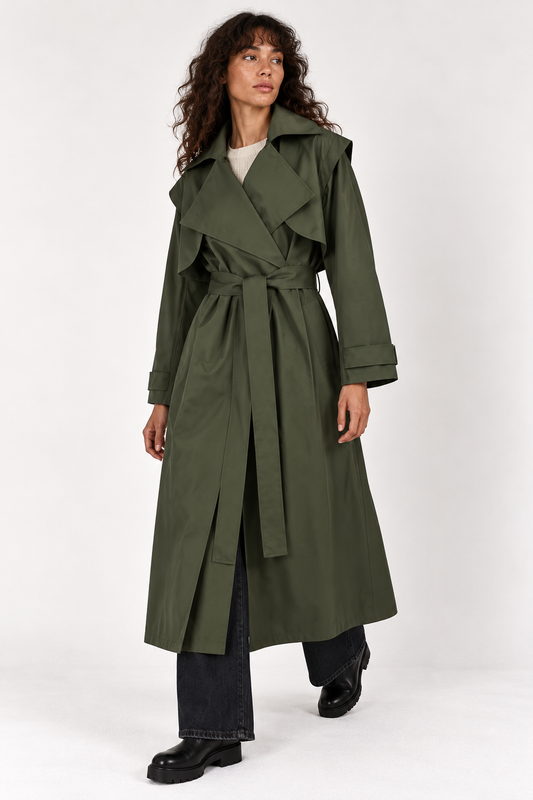 A woman modeling a long olive green trench coat with a self-tie belt, worn over a light top and dark wide-leg jeans, against a plain light gray studio background.