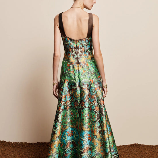 A woman stands with her back to the camera wearing a sleeveless, open-back floor-length gown with a rich green and teal floral pattern and brown straps, set against a plain beige wall and textured brown floor.