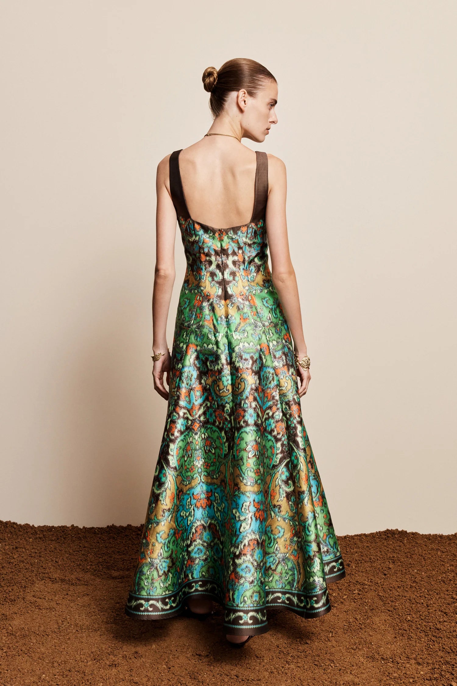 A woman stands with her back to the camera wearing a sleeveless, open-back floor-length gown with a rich green and teal floral pattern and brown straps, set against a plain beige wall and textured brown floor.