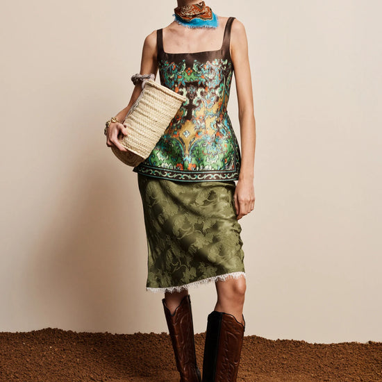 A model stands against a beige wall and brown textured floor wearing a sleeveless printed top and matching green skirt, with brown knee-high boots and a woven straw bag.