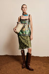 A model stands against a beige wall and brown textured floor wearing a sleeveless printed top and matching green skirt, with brown knee-high boots and a woven straw bag.