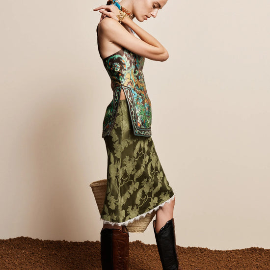 A woman models a sleeveless green midi dress with a floral pattern and a matching patterned belt, styled with brown and black knee-high boots against a beige studio backdrop.