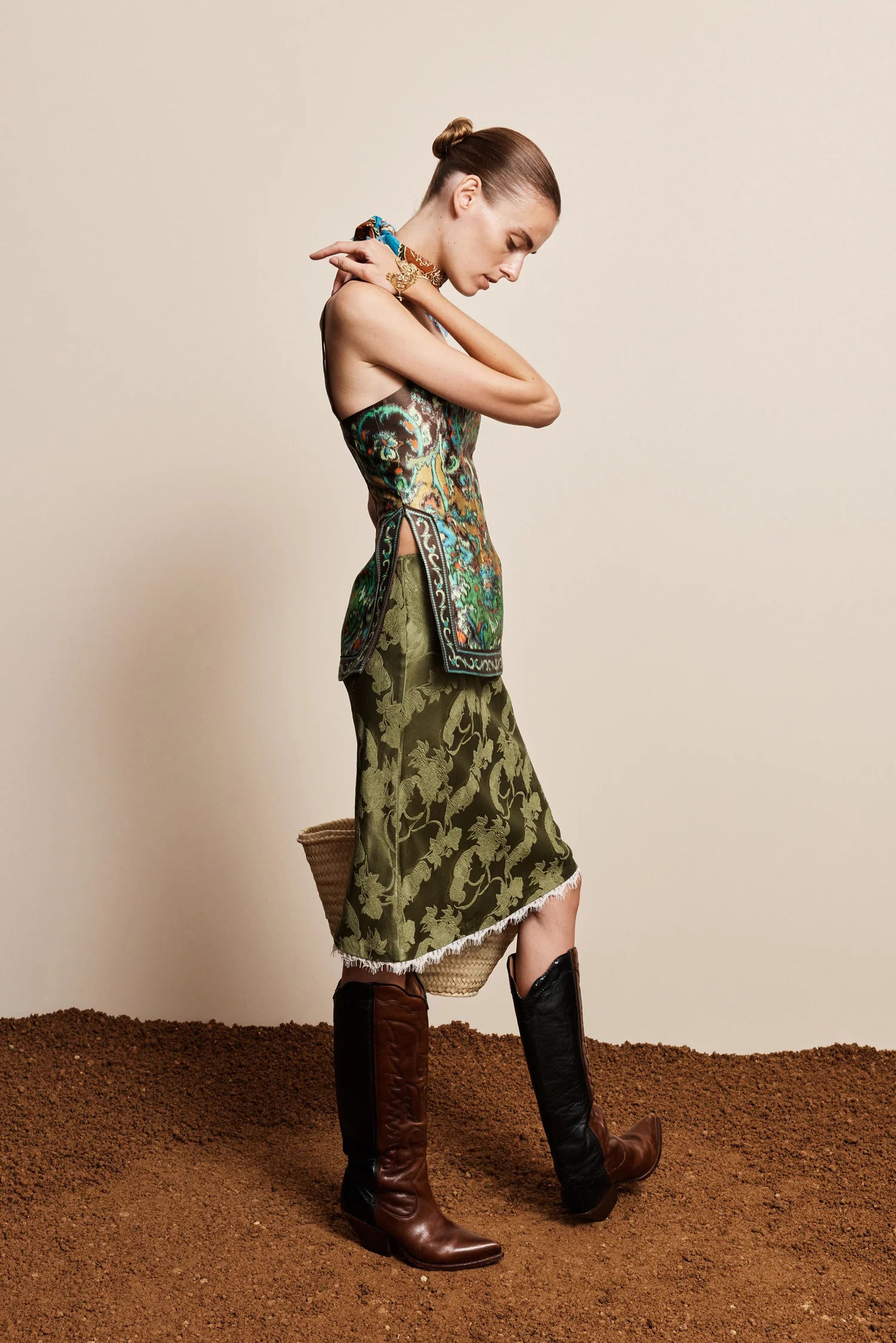 A woman models a sleeveless green midi dress with a floral pattern and a matching patterned belt, styled with brown and black knee-high boots against a beige studio backdrop.