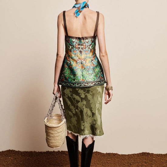 Woman wearing a floral top and green skirt with a blue scarf, standing on a textured surface.