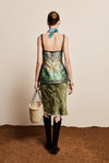 Woman wearing a floral top and green skirt with a blue scarf, standing on a textured surface.