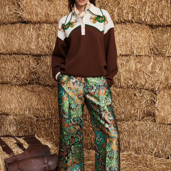 A model stands in front of stacked hay bales wearing a brown and cream color-blocked sweater with green embroidered details, patterned green and blue wide-leg trousers, and black shoes. A brown leather satchel is placed on the hay bale to her left.