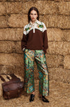 A model stands in front of stacked hay bales wearing a brown and cream color-blocked sweater with green embroidered details, patterned green and blue wide-leg trousers, and black shoes. A brown leather satchel is placed on the hay bale to her left.