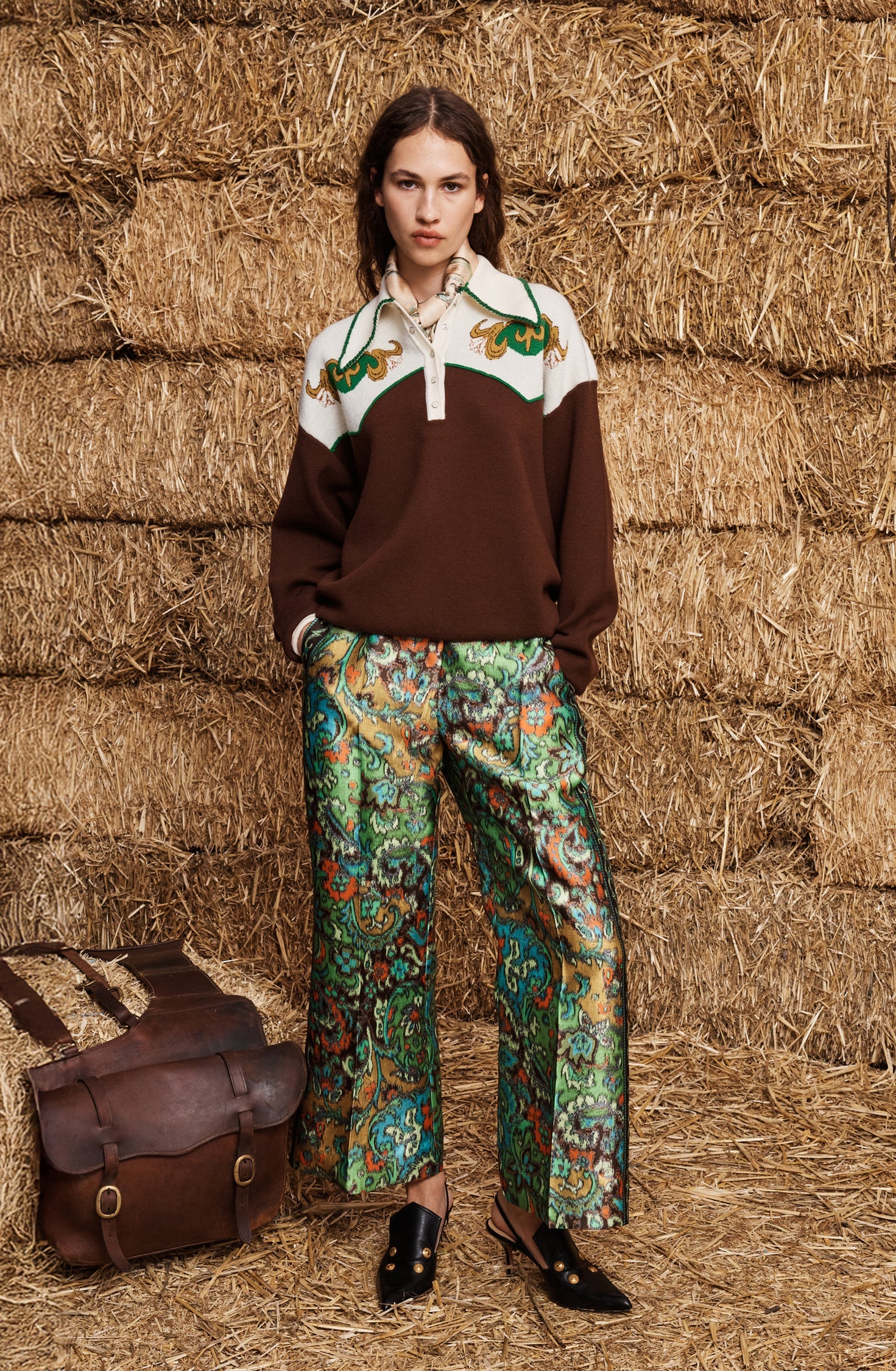 A model stands in front of stacked hay bales wearing a brown and cream color-blocked sweater with green embroidered details, patterned green and blue wide-leg trousers, and black shoes. A brown leather satchel is placed on the hay bale to her left.