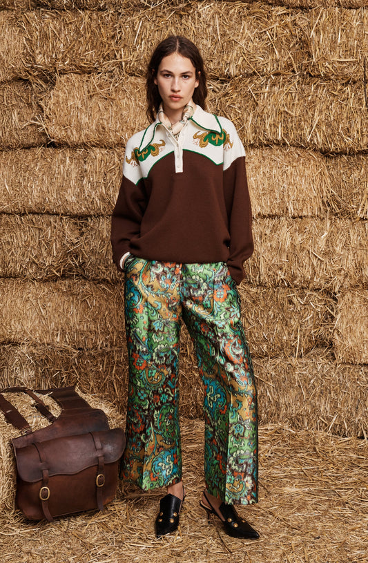 A model stands in front of stacked hay bales wearing a brown and cream color-blocked sweater with green embroidered details, patterned green and blue wide-leg trousers, and black shoes. A brown leather satchel is placed on the hay bale to her left.
