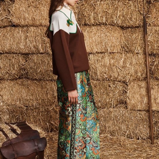 A woman stands in profile against a backdrop of stacked hay bales, wearing a brown knit sweater over a white top and a colorful floral maxi skirt. A brown leather bag rests on the straw floor beside her.