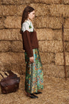A woman stands in profile against a backdrop of stacked hay bales, wearing a brown knit sweater over a white top and a colorful floral maxi skirt. A brown leather bag rests on the straw floor beside her.
