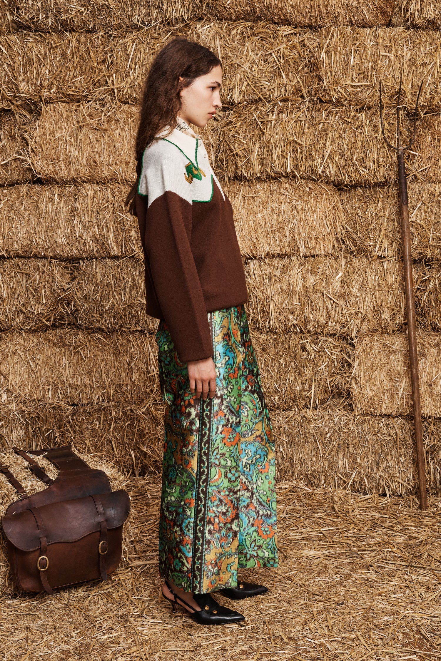 A woman stands in profile against a backdrop of stacked hay bales, wearing a brown knit sweater over a white top and a colorful floral maxi skirt. A brown leather bag rests on the straw floor beside her.