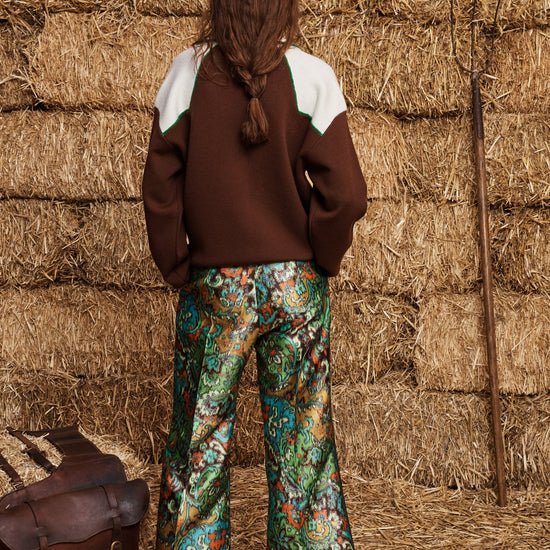 A model viewed from behind wearing a brown and white color-blocked sweater and green paisley wide-leg trousers, standing in front of a stack of hay bales with a brown leather bag at her side.