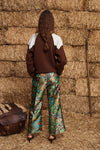A model viewed from behind wearing a brown and white color-blocked sweater and green paisley wide-leg trousers, standing in front of a stack of hay bales with a brown leather bag at her side.