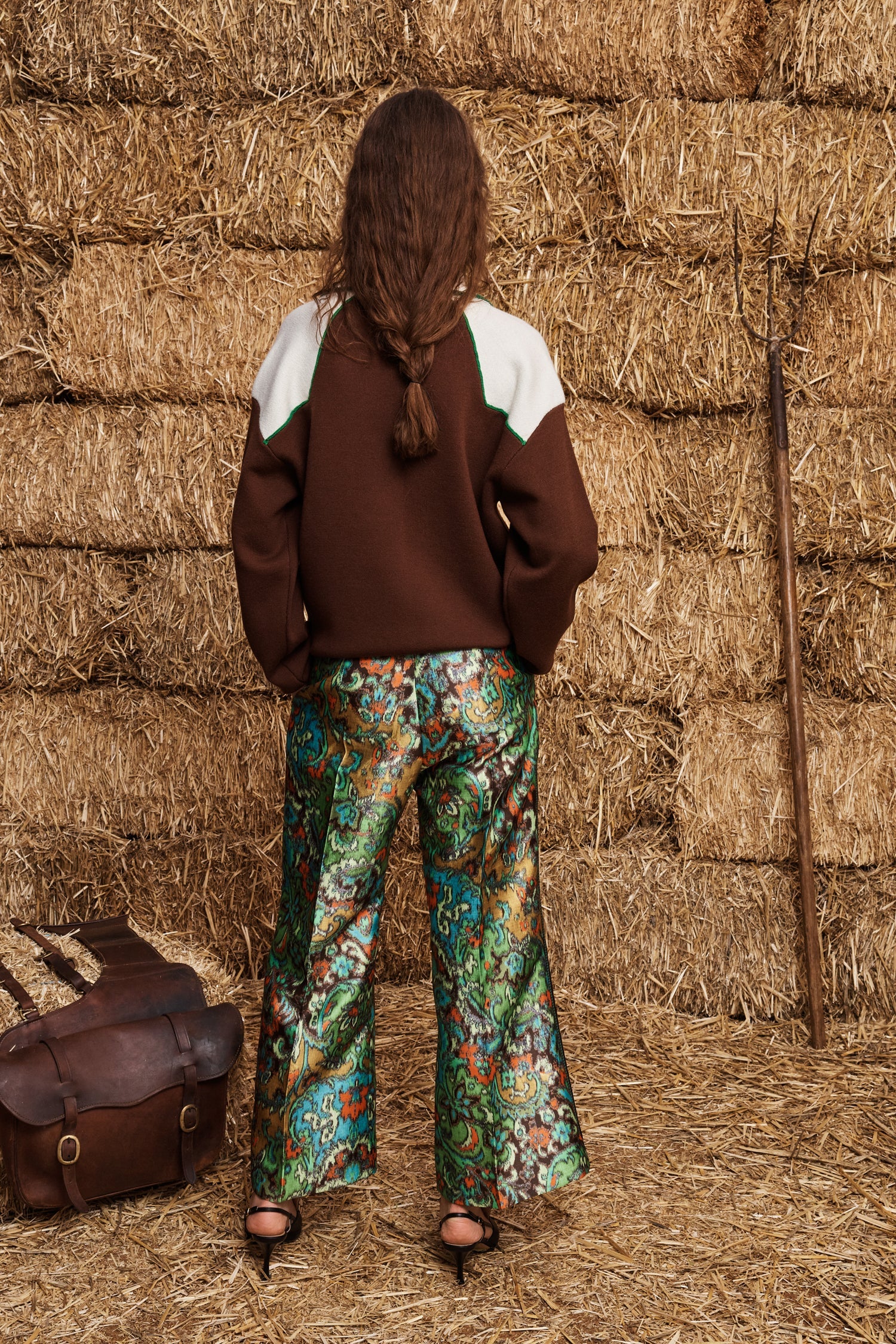 A model viewed from behind wearing a brown and white color-blocked sweater and green paisley wide-leg trousers, standing in front of a stack of hay bales with a brown leather bag at her side.