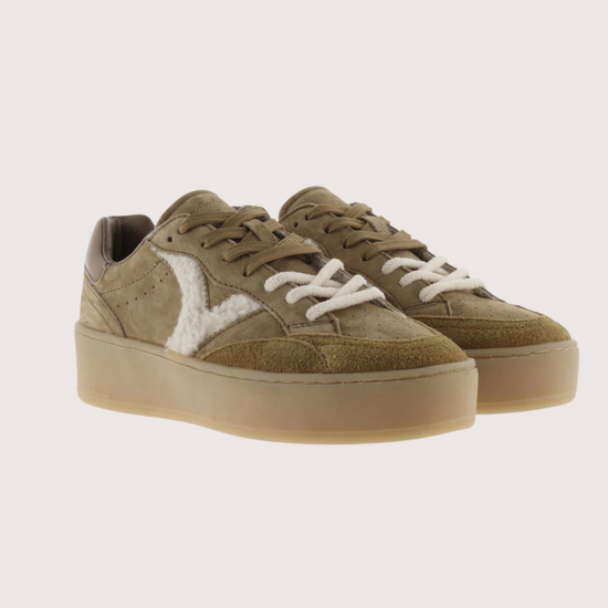 A pair of low-top platform sneakers in a muted olive-brown suede and canvas mix, featuring white laces and a white chevron logo on the side, set against a solid light beige background.