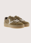 A pair of low-top platform sneakers in a muted olive-brown suede and canvas mix, featuring white laces and a white chevron logo on the side, set against a solid light beige background.