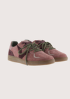 A pair of low-top suede sneakers in dusty rose and dark burgundy, featuring olive green laces and a light brown gum sole, shown against a plain light beige background.
