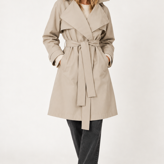 A woman modeling a beige trench coat with a matching waist tie belt, worn over a cream top and dark gray wide-leg jeans, against a plain white studio background.