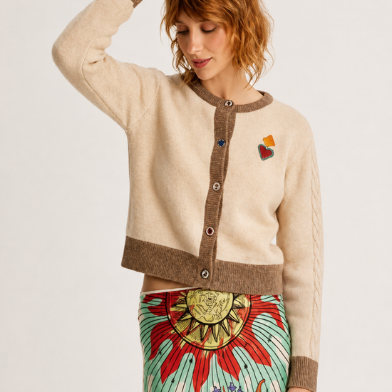 A woman modeling a cream and brown cropped cardigan sweater with a colorful embroidered heart on the chest, paired with a vibrant patterned skirt, against a plain light background.