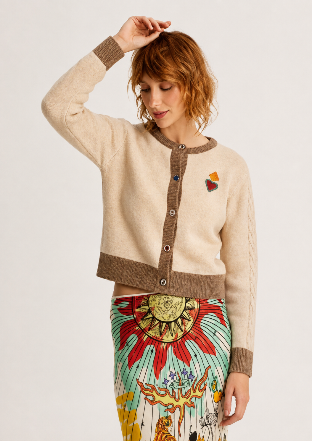 A woman modeling a cream and brown cropped cardigan sweater with a colorful embroidered heart on the chest, paired with a vibrant patterned skirt, against a plain light background.