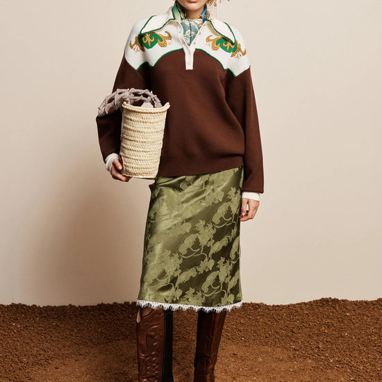 A model stands against a beige wall and brown ground, wearing a brown knit sweater with a white yoke and green floral embroidery, a green patterned skirt with white fringe, and brown knee-high boots. She holds a woven basket.