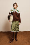 A model stands against a beige wall and brown ground, wearing a brown knit sweater with a white yoke and green floral embroidery, a green patterned skirt with white fringe, and brown knee-high boots. She holds a woven basket.